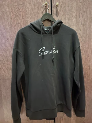 Svart hoodie med London-tryck - Svart hoodie med huva och dragsnören. På bröstet står det 'London' i vit text. Hoodien har en avslappnad passform och ribbade muddar vid ärmar och nederkant. Perfekt för en chill och stilren look.