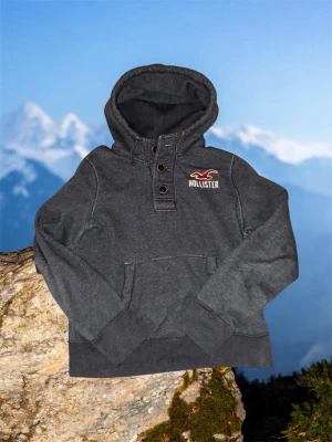 Mörkgrå hoodie från Hollister - Mörkgrå hoodie från Hollister med knappdetaljer vid halsen och klassisk magficka. Broderad logga i vitt och rött på bröstet. Mjuk och skön bomullsblandning, perfekt för chill dagar. Huva med dragsko för extra komfort. Det står att tröjan är i xl men den sitter som m/L 