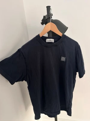 Svart t-shirt från Stone Island - Storlek M sitter som S. Snygg svart t-shirt från Stone Island med klassisk logopatch på bröstet. T-shirten har rund hals och korta ärmar, perfekt till ett avslappnat streetwear-look. Tillverkad i mjuk bomull för skön känsla hela dagen.