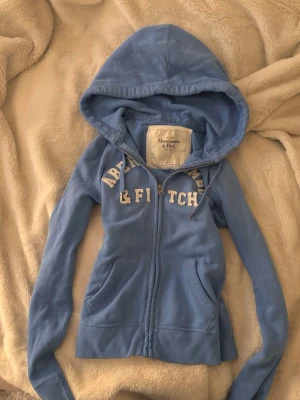 Blå kofta från Abercrombie & Fitch - Snygg ljusblå hoodie från Abercrombie & Fitch med dragkedja framtill och vita broderade bokstäver över bröstet. Hoodien har två fickor fram och justerbar huva med snören. Perfekt för en avslappnad och cool stil. Tajt passform i stl S!