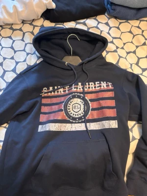 Mörkblå hoodie med tryck Saint Laurent - Snygg mörkblå hoodie med huva och magficka. Framsidan har ett stort tryck med texten 'Saint Laurent' och grafiska ränder i rött och vitt. Hoodien har dragsko i huvan och är långärmad, perfekt för en chill och avslappnad stil.