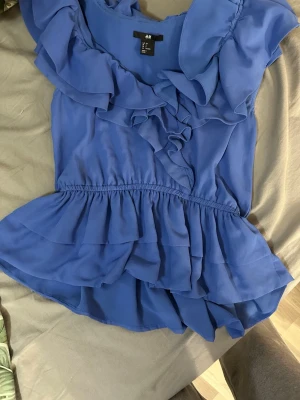 Blå volangblus från H&M - Snygg blå blus med volanger från H&M. Blusen har ett elastiskt midjeband och flera lager volanger både upptill och nedtill. Tillverkad i ett lätt, luftigt tyg som ger en flowy känsla. Perfekt för dig som gillar feminina detaljer.