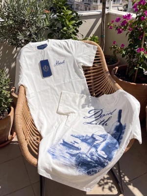 Vit t-shirt med blått tryck - Vit t-shirt med stort blått tryck på ryggen som föreställer en vinflaska, glas och strandmotiv. T-shirten har rund hals och korta ärmar, perfekt för en avslappnad stil. Materialet ser ut att vara mjuk bomull.