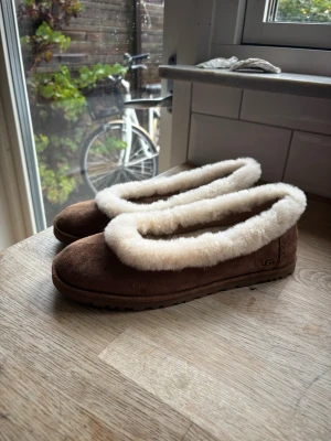 Bruna UGGS ballerina - Mysiga bruna tofflor från UGGS med fluffig vit fuskpäls runt öppningen och inuti. Tofflorna har en klassisk rundad form och är gjorda i mocka med mjuk sula. Perfekta för att hålla fötterna varma hemma. Säljer dessa i storlek 39, säljer dem för dem var för små. Om du har i vanliga fall storlek 38 så kommer dessa sitta perfekt! Aldrig använda, helt nya!