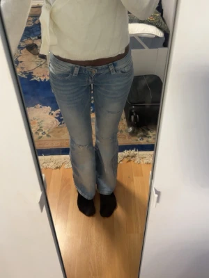 Ljusblå bootcut jeans med fickdetaljer - Säljer ett par ljusblå bootcut jeans med snygga slitningar och coola fickor med knapp bak. Jeansen har låg midja och klassisk femficksdesign. Perfekta för dig som gillar en avslappnad men trendig stil.