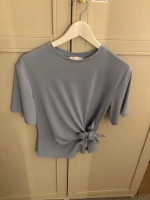 Ljusblå topp med knyt i sidan - Snygg ljusblå topp med korta ärmar och rund halsringning. Toppen har en unik detalj med knyt i sidan som ger en cool twist till din outfit. Tillverkad i ett mjukt och stretchigt material som känns skönt mot huden.