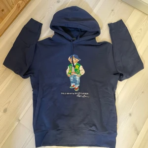 Hoodie | RALPH LAUREN. - Mörkblå hoodie med huva från Ralph Lauren, prydd med den ikoniska Polo Bear-figuren på bröstet. Trycket visar björnen i jeans, sneakers och en grön jacka. Hoodien har dragsko i huvan och ribbade muddar. Perfekt för en avslappnad och cool stil.