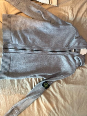 Grå ziptröja från Stone Island - Snygg grå ziptröja från Stone Island med ribbade muddar och klassisk logotyp på ärmen. Tröjan har dragkedja framtill och hög krage, perfekt för lager-på-lager. Enkel och stilren design som passar till det mesta.