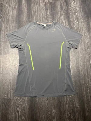 Grå tränings-tshirt med neondetaljer 2015 - Gymshark Performance T-Shirt i grått med neongröna detaljer. Äldre Gymshark-design med klassisk atletisk passform, raglanärmar och tekniskt, ventilerande material utvecklat för träning. Kontrasterande neongröna linjer framtill och den klassiska Gymshark-loggan på bröstet ger en tydlig old-school Gymshark-look. Perfekt för gym, löpning och annan träning.