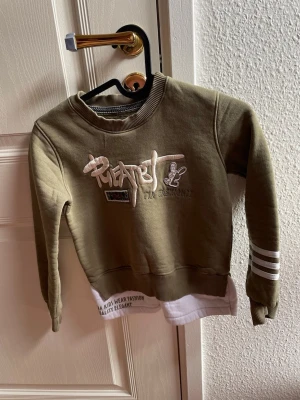 Olivgrön sweatshirt med tryck, storlek 134/140 - Cool olivgrön sweatshirt med vitt tryck och broderi framtill samt vita ränder på ena ärmen. Tröjan har ribbade muddar och en vit detalj nedtill som ger lager-på-lager känsla. Perfekt för en avslappnad och trendig stil.Finns även joggingbyxor till bland mina annonser för ett helt set.