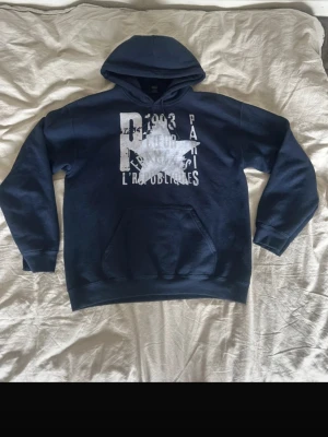 Mörkblå hoodie med tryck - Snygg mörkblå hoodie med stor vit grafisk print framtill och textdetaljer. Klassisk känguruficka och huva. Perfekt för en avslappnad och cool stil. Mjuk bomullskänsla och ribbade muddar vid ärmar och nederkant.