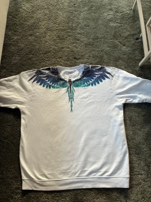 Vit sweatshirt från Marcelo burlon - Säljer en vit sweatshirt från Marcelo Burlon med ikoniskt blått och svart vingtryck över axlar och bröst. Tröjan har rund halsringning och är tillverkad i mjukt bomullsmaterial. Perfekt för dig som vill sticka ut med en unik design.