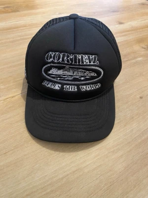Svart truckerkeps Corteiz  - Säljer en svart truckerkeps med vit brodyr framtill där det står 'CORTEIZ RULES THE WORLD' och ett broderat skepp. Sidan har vita stjärnor broderade och baksidan är i mesh för extra ventilation. Justerbar storlek med klassisk böjd skärm.