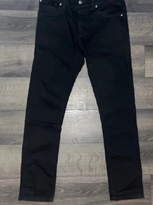 Svarta nudie jeans - Säljer ett par svarta jeans med klassisk femficksdesign och bra passform. Jeansen har knappgylf och silverfärgade nitar. Perfekta för dig som gillar en stilren och enkel look. Materialet är bomull. W32 L32