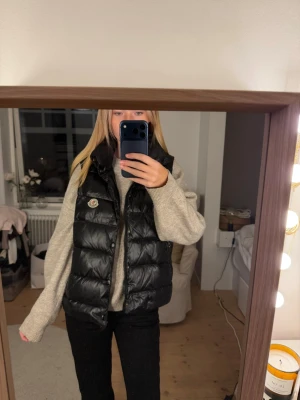 Svart dunväst från Moncler - Snygg svart dunväst från Moncler med glansig finish och klassisk logga på bröstet. Västen har hög krage och stängs med knappar och dragkedja. Perfekt att slänga över en hoodie eller stickad tröja för en cool och avslappnad look.