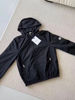 Svart vindjacka med huva - Snygg svart vindjacka från Moncler med huva och dragkedja framtill. Jackan har två fickor med dragkedja, elastiska muddar och en Moncler-logga på ärmen. Perfekt för blåsiga dagar och enkel att matcha till olika outfits.