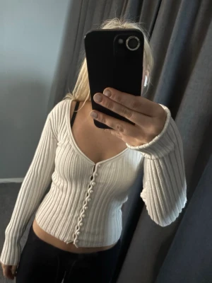 Vit ribbad kofta från NA-KD - Säljer en vit ribbad kofta från NA-KD med långa ärmar och djup v-ringning. Koftan har pärlknappar framtill och är figurnära i modellen. Perfekt att styla med både jeans och kjol för en trendig look.