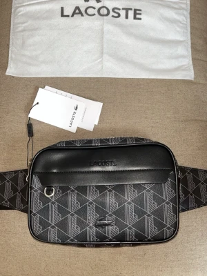 Lacoste Väska - Säljer en  helt ny svart grå Lacoste midje väska. Jättefin och bra för o hålla saker, passar till mycket, kommer med alla förpackningar!!  —- PRISET KAN DISKUTERAS VID SNABB AFFÄR!!