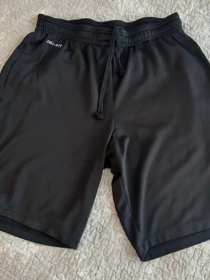 Nike  Fri Fit Svarta träningsshorts M  - Snygga svarta träningsshorts med elastisk midja och snörning. Tillverkade i ett lätt och snabbtorkande material, perfekta för gymmet eller löprundan. DRI-FIT-tryck på sidan ger en sportig look. Klassisk passform och enkel design.