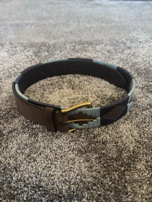 Brunt och blått mönstrat bälte - Snyggt bälte i brunt skinn med blått och beige geometriskt mönster. Bältet har en metallspänne i mässingsfärg och detaljer i olika blå nyanser. Perfekt för att lyfta din outfit med en unik accessoar.