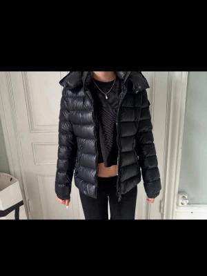 Moncler dunjacka  - Snygg svart dunjacka från Moncler med glansig finish och puffig design. Jackan har huva, dragkedja framtill och Moncler-logga på ärmen. Perfekt för kalla vinterdagar och ger en trendig look. Kontakta gärna vid frågor och innan köp!💕