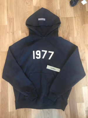 Svart Essentials Fear of God - Snygg svart hoodie från Essentials Fear of God med trycket '1977' på bröstet. Klassisk oversized passform, stor huva och magficka framtill. Tillverkad i mjukt bomullsmaterial som är perfekt för en avslappnad streetwear-look.