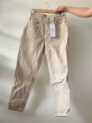 Beige mom jeans Dagny - Säljer ett par beige mom jeans från Gina tricot, modell Dagny i storlek 34. Jeansen har hög midja och raka ben.  Oanvända med prislapp kvar.