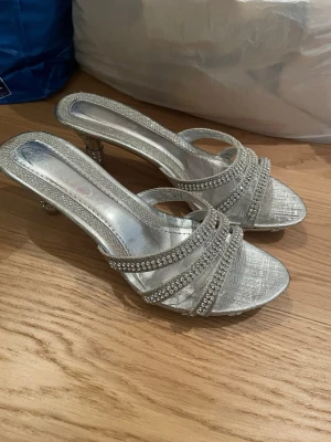 Silvriga kitten heels med strassdetaljer - Säljer ett par glittriga silvriga kittens med öppen tå och smala remmar dekorerade med strass. Skorna har en låg klack och en elegant, festlig design. Perfekta för att lyfta vilken outfit som helst med extra bling.