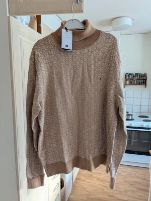 Beige stickad polotröja Tommy Hilfiger - Snygg beige stickad polotröja från Tommy Hilfiger med diskret diamantmönster i vitt. Tröjan har ribbade muddar och hög krage i en matchande beige ton. Liten broderad logga på bröstet. Perfekt för dig som gillar klassisk och stilren design. Storlek XXXL 