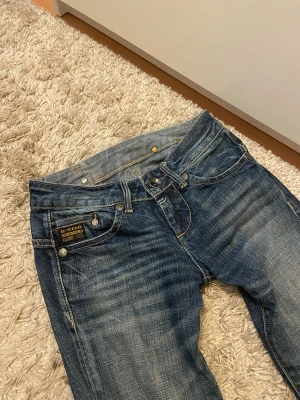 Blå bootcut jeans från G-Star - Snygga blå jeans från G-Star med bootcut passform och slitna detaljer. De är lågmidjade men inte supermycket. Innerbenslängd: ca 75 cm. Midjemått rakt över: 32 cm. Hör av dig vid frågor eller intresse💕