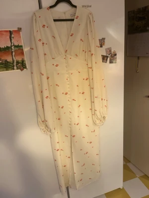Lång vit klänning med blommor - Romantisk långklänning i vit ton med små röda blommor, v-ringad framtill och långa puffärmar. Klänningen har knappar längs hela framsidan och är tillverkad i ett lätt tyg, perfekt för sommarens varma dagar.