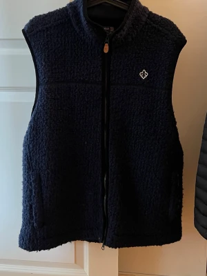 Morris Pälsväst - Mysig mörkblå fleeceväst med hög krage och hel dragkedja framtill. Västen har två fickor och en liten vit broderad logga på bröstet. Perfekt för lager-på-lager och håller dig varm under kyliga dagar.