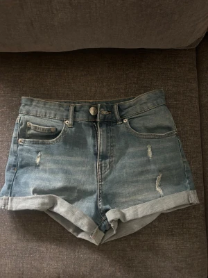 Blå denimshorts med upprullad kant - Snygga blå denimshorts med upprullad kant och slitna detaljer framtill och baktill. Klassisk femficksmodell med knapp och dragkedja i midjan. Perfekta för varma dagar och enkel att matcha med olika toppar.