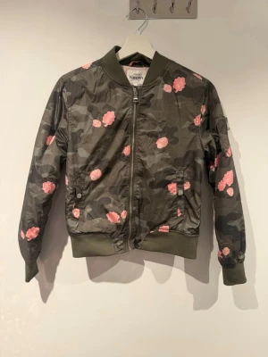 Camo bomberjacka med rosa blommor - Snygg bomberjacka i camomönster med rosa blommor. Jackan har ribbade muddar, dragkedja framtill och fickor med knapp. Färgen är militärgrön med rosa detaljer och materialet är 100% polyester. Perfekt nu till hösten. 