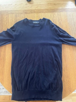 Mörkblå stickad tröja Dressmann - En stilren mörkblå stickad tröja från Dressmann i Supima-bomull. Tröjan har rund halsringning, långa ärmar och ribbade muddar vid ärmslut och nederkant. Perfekt för en clean och enkel look. Nästan oanvänd