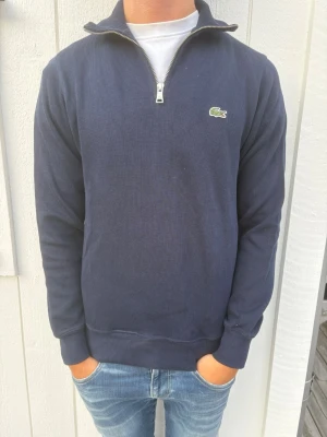 Mörkblå halvzip tröja från Lacoste - Snygg mörkblå tröja från Lacoste med halv dragkedja och klassisk krokodillogo på bröstet. Tröjan har ribbade muddar och krage, perfekt för en clean och stilren look. Passar till jeans eller chinos för en avslappnad men ändå uppklädd stil.