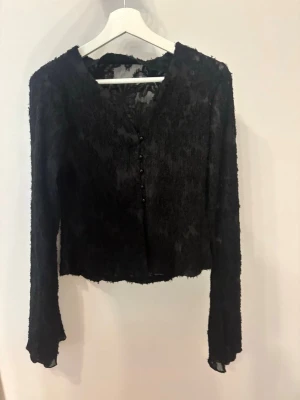 Svart transparent blus med knappar - Snygg svart blus i transparent mesh med struktur och dekorativa knappar framtill. Blusen har lång ärm och råa kanter vid ärmslut och nederkant. Perfekt för dig som gillar en edgy och trendig stil.