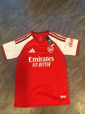 Arsenal matchtröja röd/vit Adidas 2026/2027 - Snygg Arsenal matchtröja från Adidas i rött och vitt med klubbmärke och sponsortryck på bröstet. Tröjan har korta ärmar, rund hals och klassiska Adidas-ränder på axlarna. Tillverkad i lätt och ventilerande material, perfekt för fotboll eller träning.