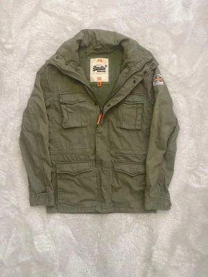 Superdry Jacka S - Tja! Säljer nu denna riktigt feta Superdry Jacka i storlek S. Perfekt till hösten/våren. Den är i mycket fint skick, bortsett från att dragkedjan strular. Den har fullt fungerande knappar hela vägen så det påverkar inte användning eller utseendet alls. Vid eventuella frågor eller funderingar är det bara att höra av sig! 