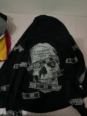 Svart Philipp Plein hoodie med dödskalle - Säljer en svart hoodie från Philipp Plein med dragkedja och huva. Hoodien har ett stort dödskallemotiv på ryggen och vita logoband med texten 'PHILIPP PLEIN 1978' över hela plagget. Materialet är mjukt och bekvämt, perfekt för en streetwear-look.