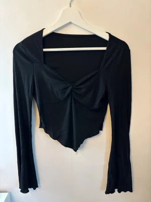 Svart långärmad croppad topp - Snygg svart långärmad topp med croppad passform och twistad detalj framtill. Toppen har en asymmetrisk nederkant och är tillverkad i ett mjukt, stretchigt material som sitter bekvämt på kroppen. Perfekt för dig som gillar trendiga och stilrena plagg.