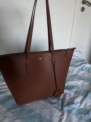 Brun toteväska från Ralph Lauren - Snygg brun toteväska från Ralph Lauren i strukturerat konstläder. Väskan har långa axelband, guldfärgade detaljer och en dekorativ tag med RL-logga. Perfekt för dig som vill ha plats för allt du behöver och samtidigt hålla stilen.