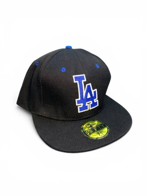 Svart LA Dodgers keps - Snygg svart keps med blå LA Dodgers-logga framtill och blå detaljer. Klassisk flat brim-modell med broderade ventilationshål och MLB-logga bak. Tillverkad i ett slitstarkt tyg som ger en clean och sportig look.