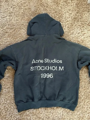 Mörkgrå hoodie Acne Studios - Snygg mörkgrå hoodie från Acne Studios med stor huva och tryck på ryggen där det står 'Acne Studios STOCKHOLM 1996' i vitt. Hoodien har en avslappnad passform och är tillverkad i mjukt bomullsmaterial. Perfekt för en casual streetwear-look.