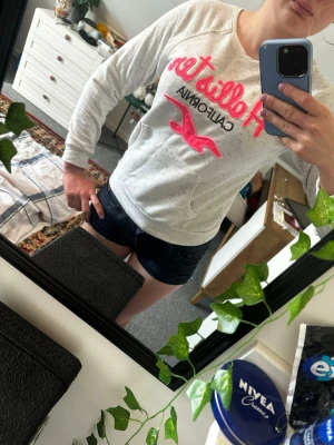 Vit Hollister hoodie med rosa logga - Säljer en vit hoodie från Hollister med gråmelerade detaljer och stor rosa Hollister-logga samt texten 'California' på bröstet. Hoodien har en magficka och långa ärmar med ribbade muddar. Perfekt för en avslappnad och trendig look.