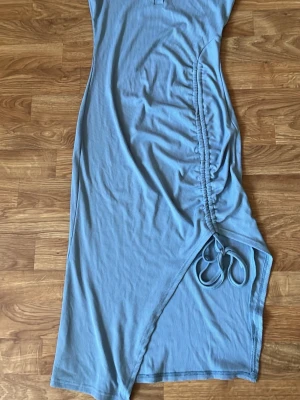 Blå ribbad långklänning med snörning - Snygg blå långklänning i ribbad bomull med ärmlös design. Klänningen har knappar framtill vid halsen och en cool snörningsdetalj på sidan som ger en asymmetrisk look. Perfekt för dig som gillar trendiga detaljer och bekväma material.