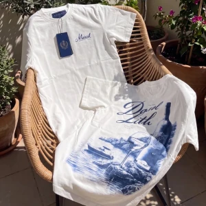 Vit t-shirt med blått tryck - Säljer en vit t-shirt med blått tryck på ryggen som visar en vinflaska, glas och havsmotiv. Framtill finns en liten blå logga/text på bröstet. Klassisk passform och rund hals. Perfekt för dig som gillar stilrena plagg med unik detalj.