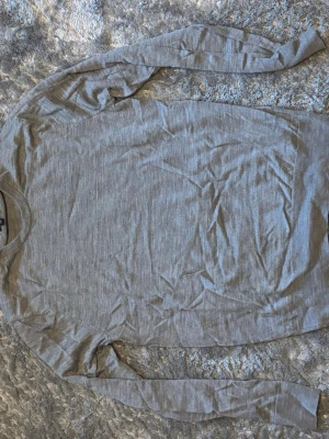 Ljusgrå långärmad tröja - En stilren ljusgrå långärmad tröja i tunt, mjukt material. Tröjan har rund halsringning och en enkel, klassisk design som passar till det mesta. Perfekt för lager på lager eller som basplagg i garderoben.