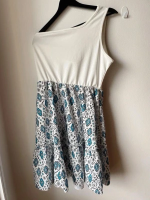 Vit oneshoulder klänning med mönster - Säljer en snygg oneshoulder klänning med vit överdel och mönstrad kjol i blått och svart. Kjolen har ett grafiskt mönster och klänningen är kort i modellen. Perfekt för sommarens varma dagar!