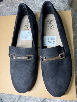 Svarta loafers med metallspänne - Säljer ett par svarta loafers i mockaimitation med snyggt metallspänne framtill. Skorna har rundad tå och låg klack, perfekta för en stilren look. Märket är So All och storleken är 39.